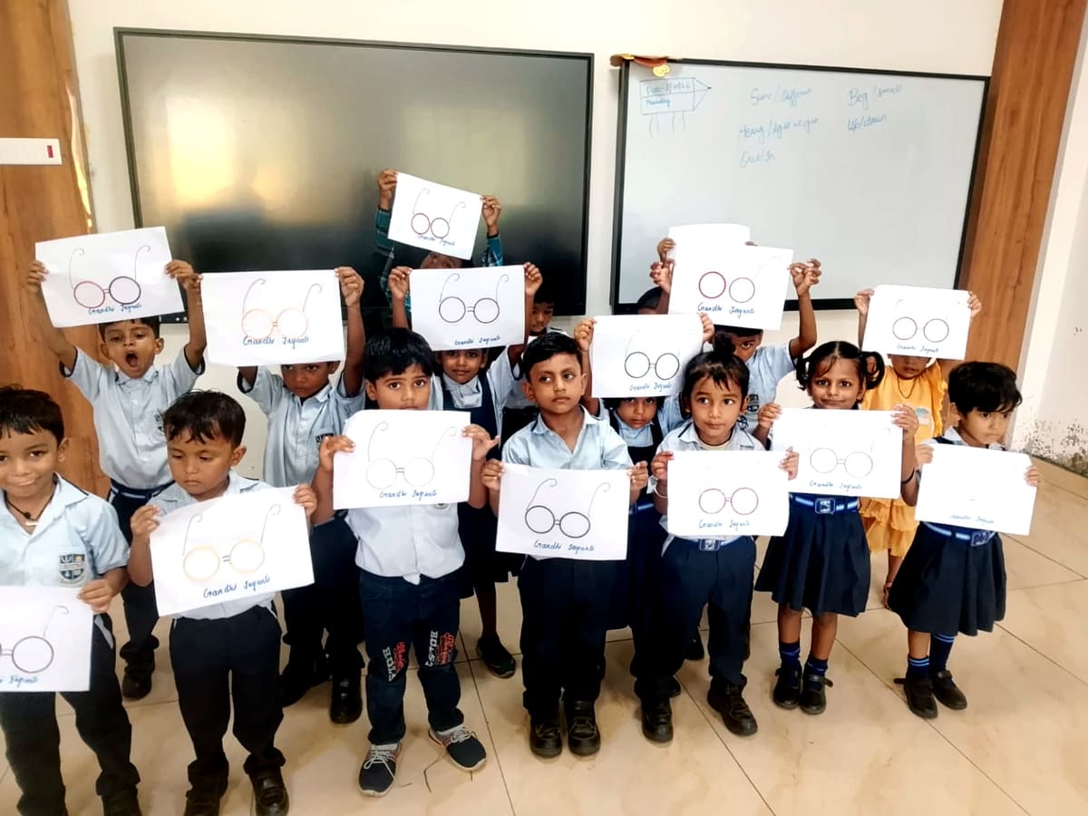 Pre-primary pupils in school uniform hold up drawings of Gandhi's round spectacles in a classroom with an interactive display panel.