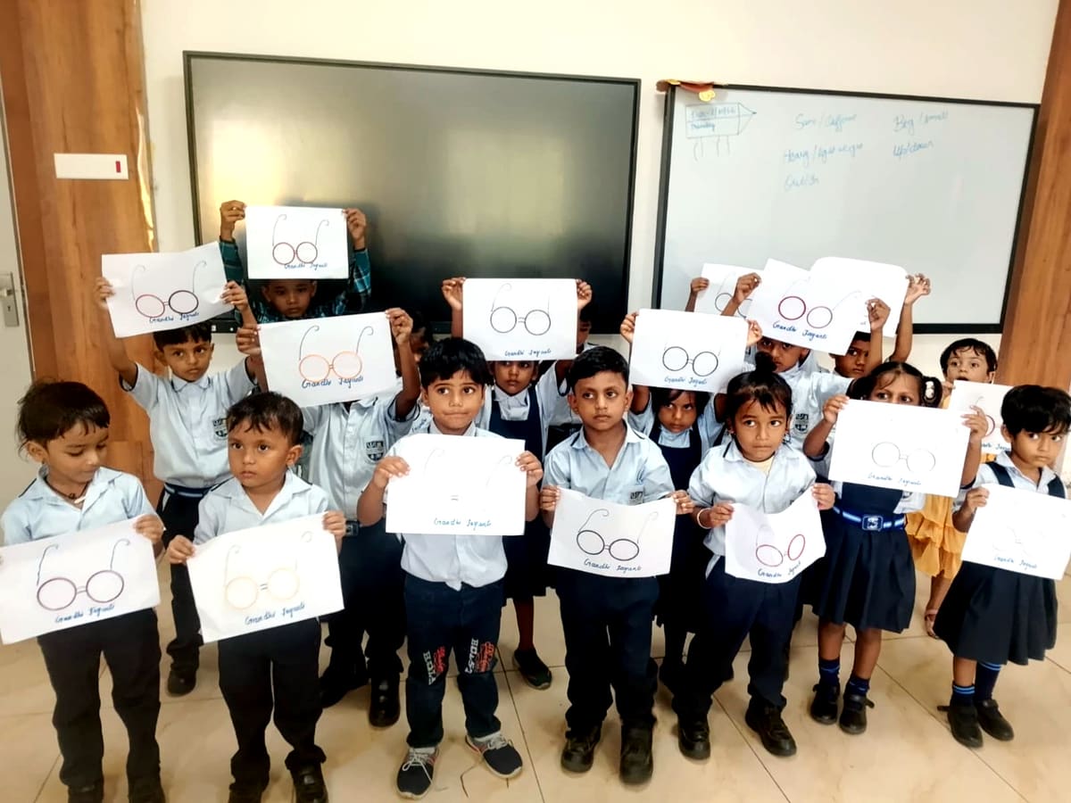 A class of young pupils stands together holding their Gandhi Jayanti spectacles artwork.
