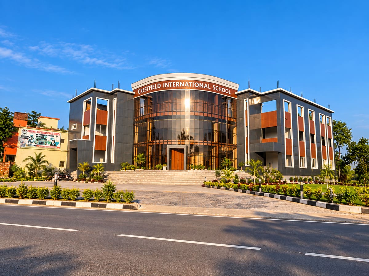 The Crestfield International School building: a three-storey façade with a curved glass entrance under the school's name, steps and landscaped gardens, seen from the road.